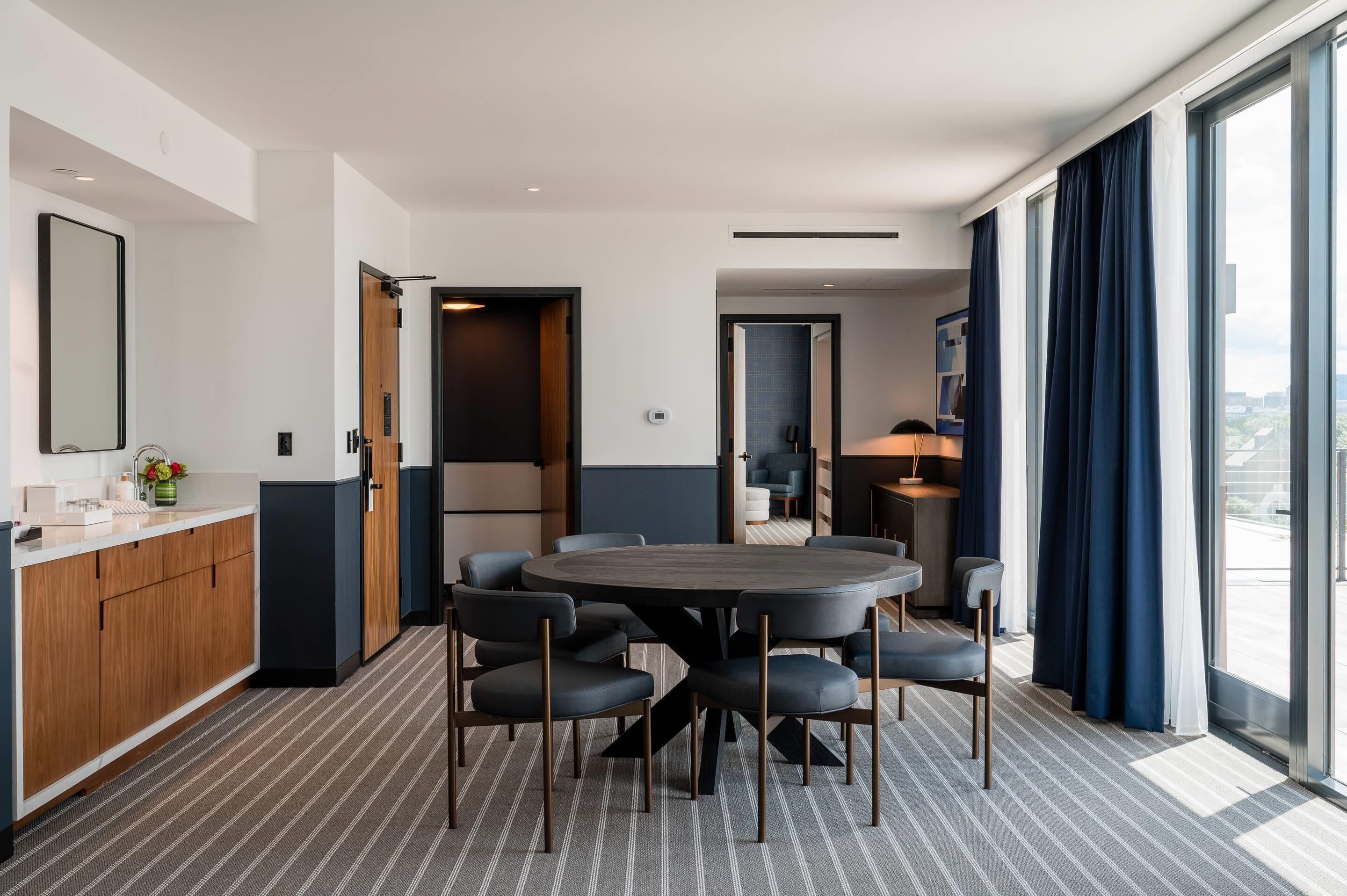 A modern hotel room with a dining area, a kitchen, and a large window with curtains.