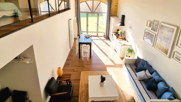 A spacious loft with a high ceiling, exposed wooden beams, a dining area with a table and chairs, a sofa with cushions, and a view through a large window. - Loft in the heart of the vineyards in a former Chai dating from 1815 (Marcy)
