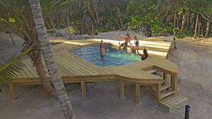 Um deck de madeira ao redor de uma piscina, com uma praia de areia e palmeiras ao fundo.
