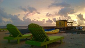 Duas poltronas verdes numa praia arenosa, com um posto de salva-vidas e barcos ao fundo.