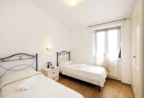 A bedroom with two beds, a nightstand, a window with sheer curtains, and a wall-mounted light fixture. - Detached  Villa, 55 square meters, on the ground floor with private garden of 150 square meters with barbecue and sunbeds (Vieste)