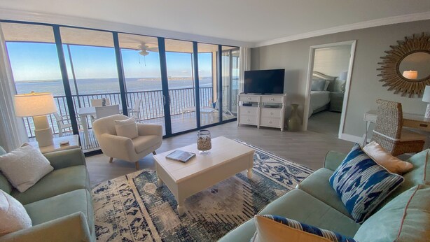 Un vaste salon doté d’une grande porte-fenêtre coulissante donnant sur un balcon avec vue sur la mer. On y trouve un téléviseur posé sur un meuble blanc, une table basse et un canapé agrémenté de coussins à motifs.
