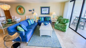 Un salon avec un canapé bleu, un fauteuil vert, une table basse blanche et une table à manger en verre, accompagnée de chaises.