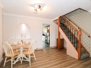 A dining area with a round table and four chairs, a kitchen visible through an open doorway, and a wooden staircase with a patterned railing. - 57 Southend Road (Hunstanton)