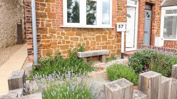Ein Steinhäuschen mit Hausnummer 57, einer hölzernen Bank und einem Garten mit Lavendel und anderen Pflanzen.