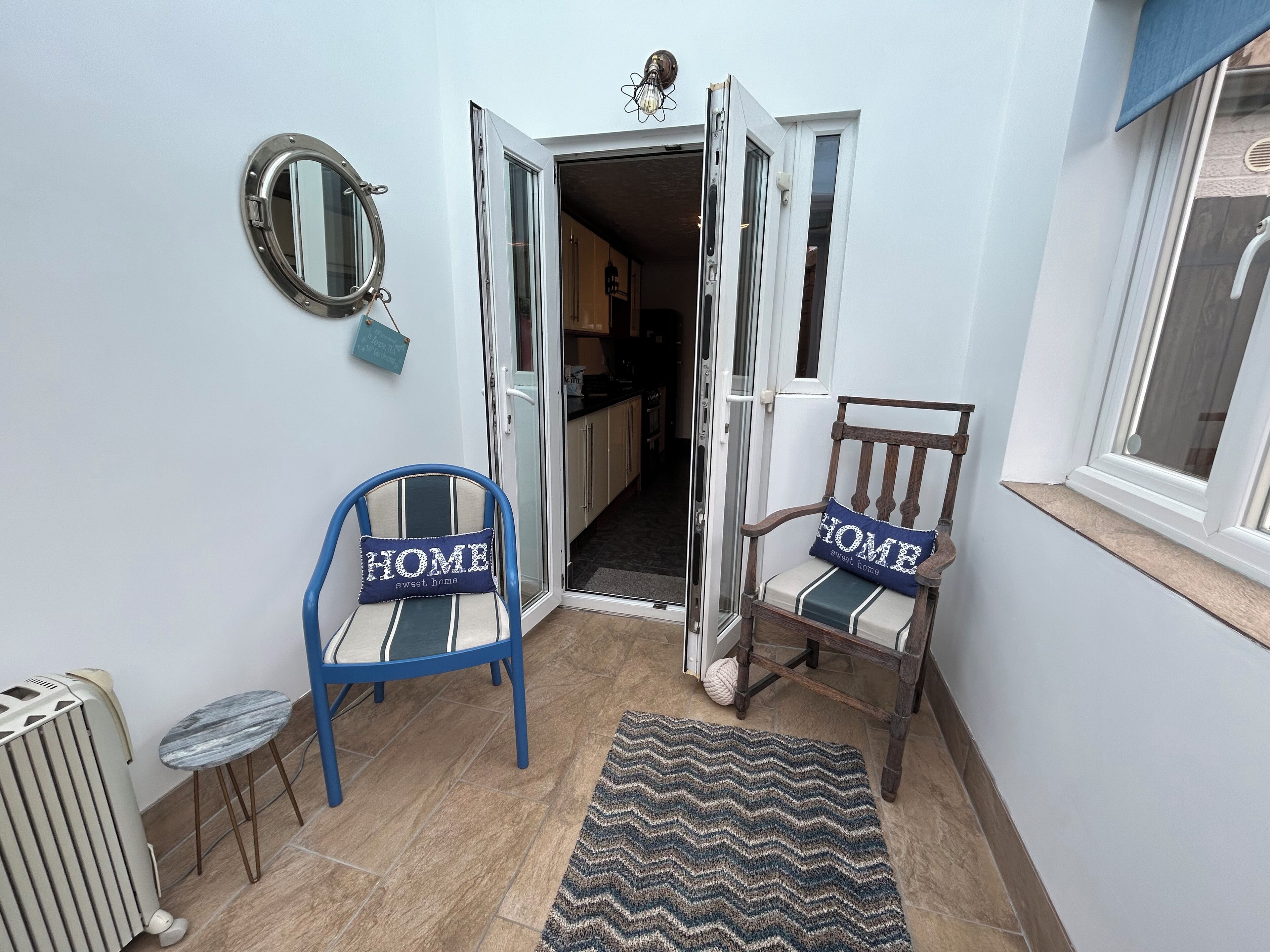 A narrow hallway with two chairs, each with a 'HOME sweet home' pillow, a small table, a radiator, and a rug leading to an open door.