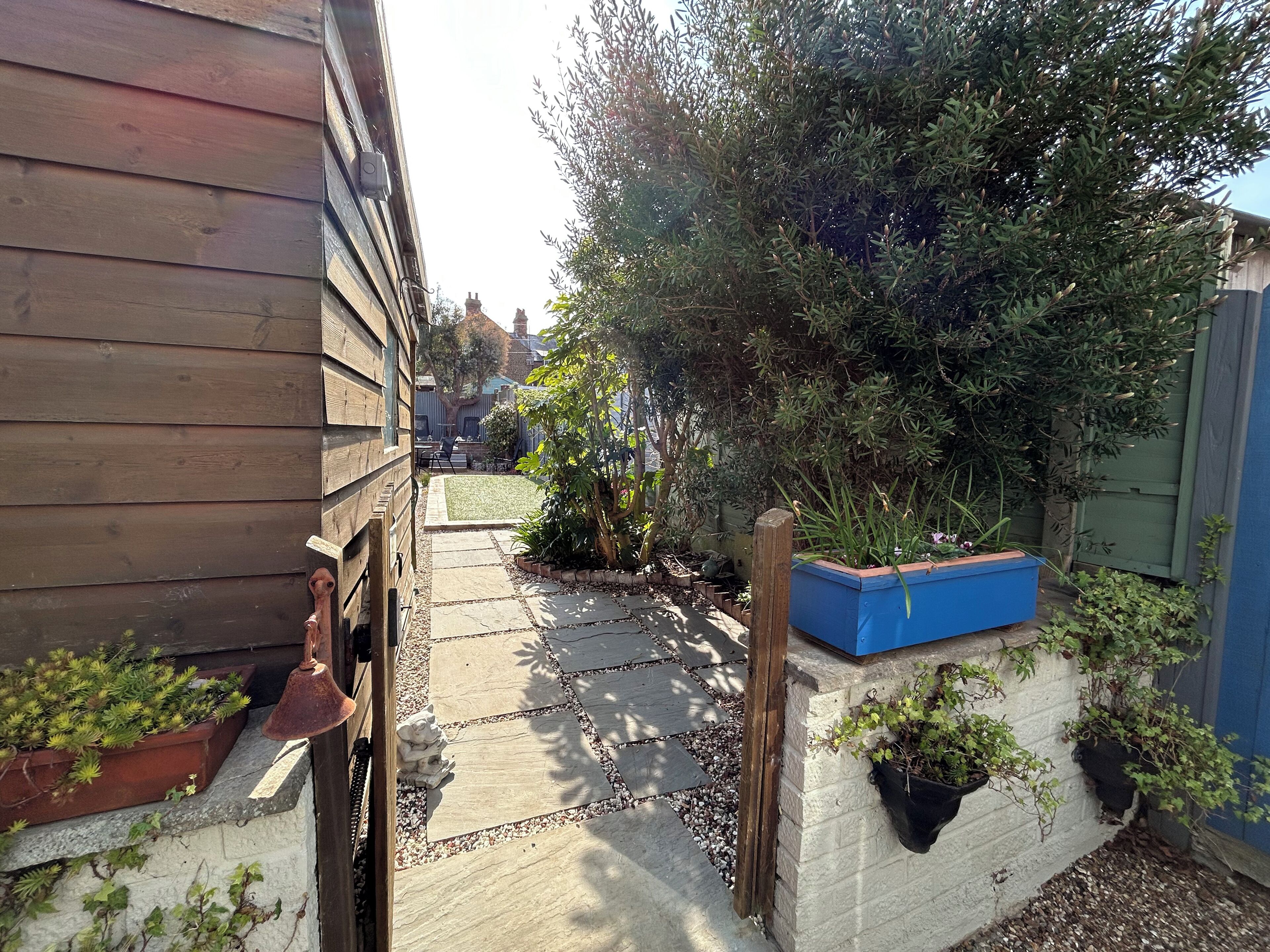 A narrow pathway with a stone pavement leading to a wooden building. There are potted plants and a blue planter box along the path.