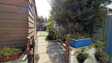A narrow pathway with a stone pavement leading to a wooden building. There are potted plants and a blue planter box along the path.