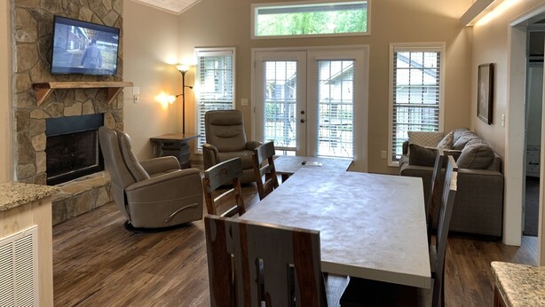 A spacious living room with a stone fireplace, comfortable seating, a dining table, and large windows allowing natural light.