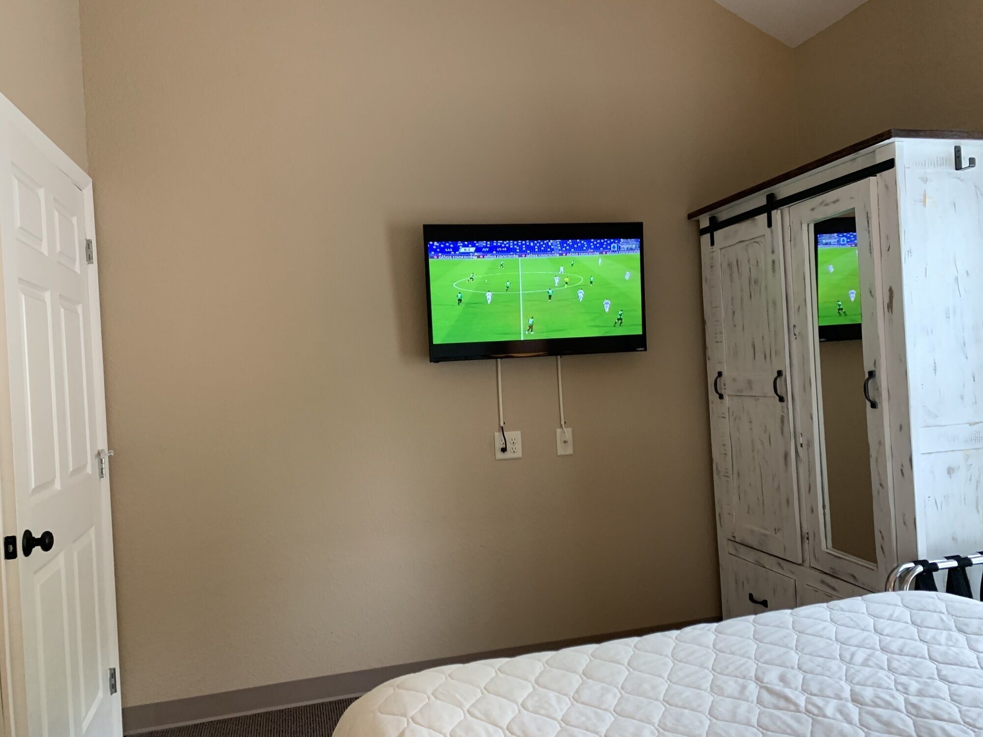 Une chambre à coucher avec un lit, un téléviseur fixé au mur qui diffuse un match de football, et une armoire.
