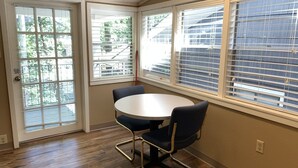 Ein Zimmer mit einem runden Tisch, zwei Stühlen, großen Fenstern mit Jalousien und einer Tür mit Blick nach draußen.