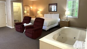 Ein Hotelzimmer mit einem Bett, zwei Sesseln, einem kleinen Tisch, einem Nachttisch, einer großen Badewanne und einem Fenster mit Jalousien.