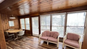 Una habitación con paneles de madera, una mesa de comedor y sillas, dos sofás estampados y ventanas grandes con persianas.