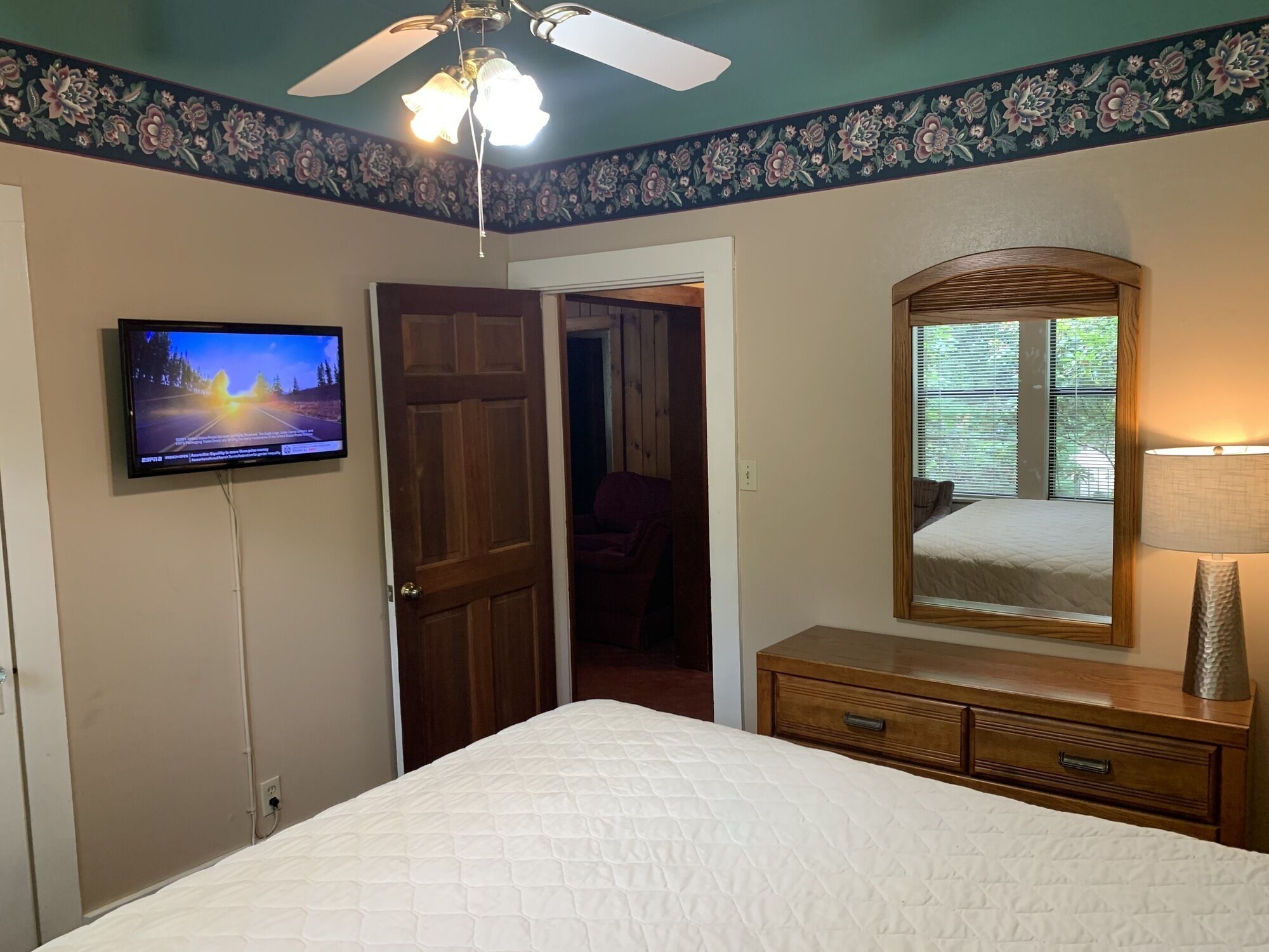 A bedroom with a bed, a television mounted on the wall, a ceiling fan, a dresser, and a window with blinds.
