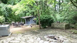 Un patio de piedra con un pequeño hogar, una terraza de madera y una estructura tipo cabaña azul rodeada de árboles.
