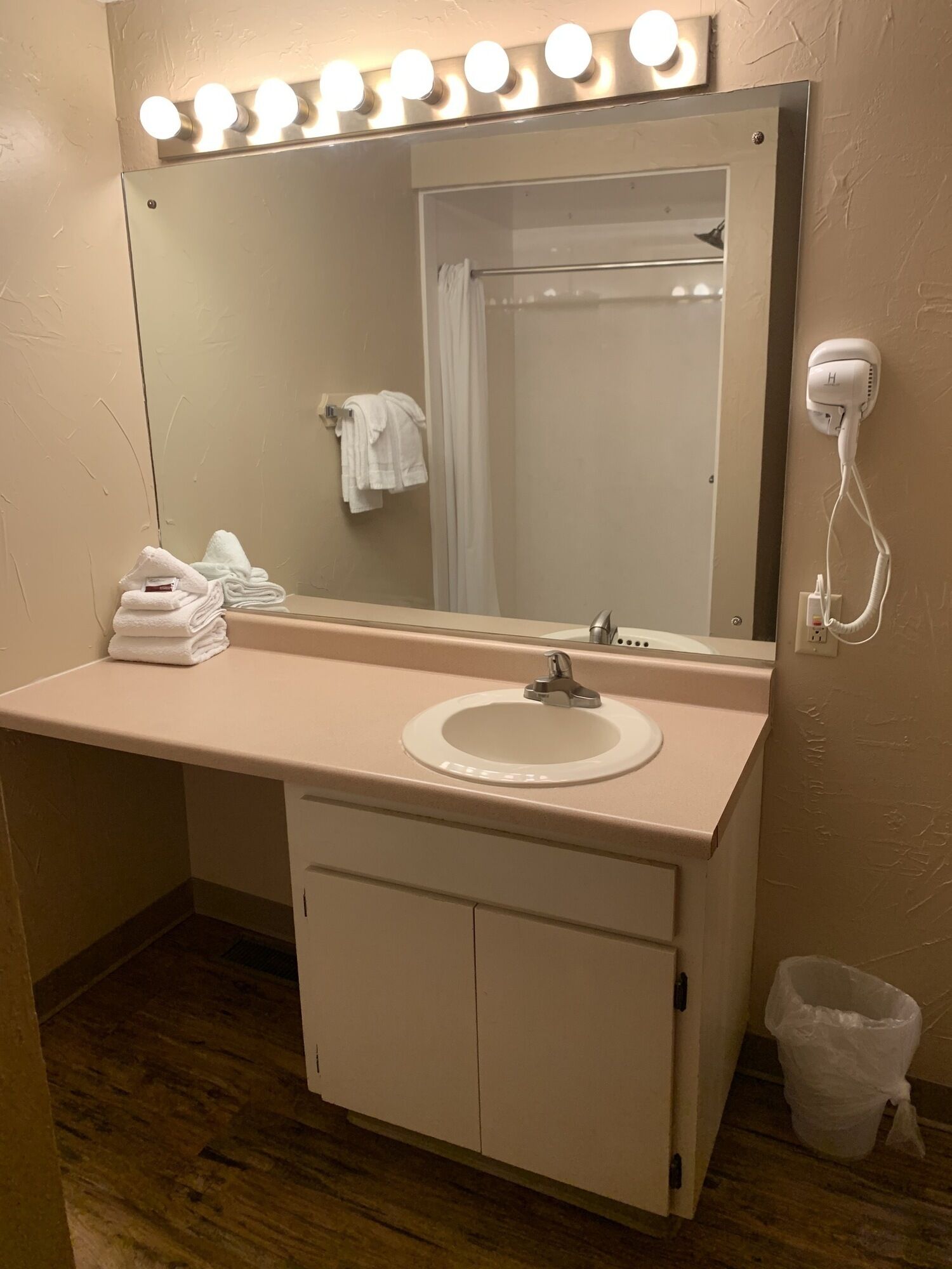 A bathroom with a vanity, sink, and mirror. There are towels stacked on the vanity, a hairdryer on the wall, and a trash can.