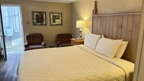 Una habitación de hotel con una cama grande, dos sillones, una mesita de noche con una lámpara y dos cuadros en la pared.
