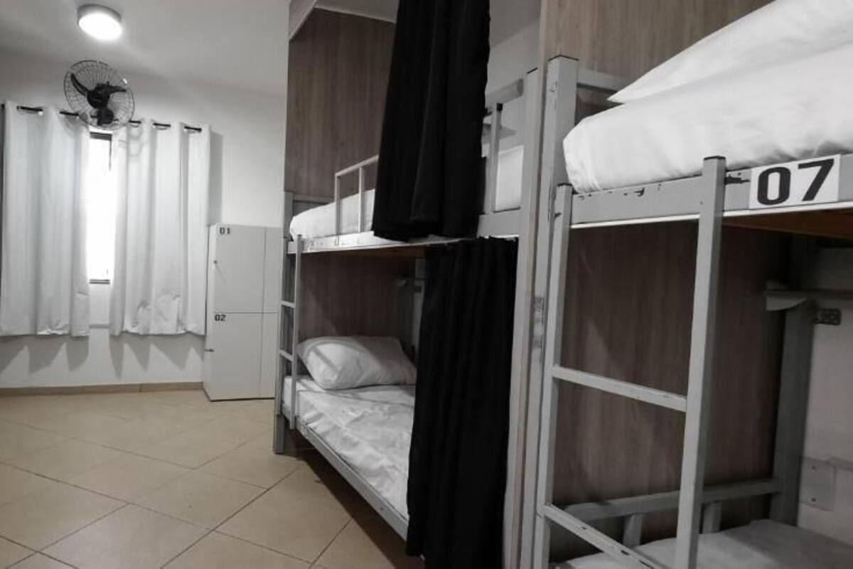 Quarto tipo dormitório com beliches, ventilador de teto e janela com cortinas.