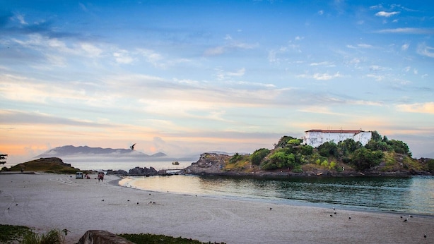 A coastal scene with a white building on a small island, a sandy beach, and calm waters. - Soul do Mar Hostel (Cabo Frio)