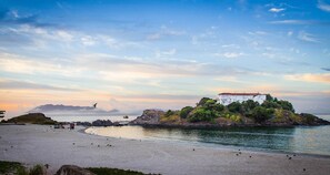Un paisaje costero con un edificio blanco en una pequeña isla, una playa arenosa y aguas tranquilas.