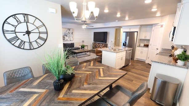Een moderne keuken met een centraal eiland, een eetgedeelte met een grote houten tafel, en een woongedeelte met een bank en een grote wandklok.