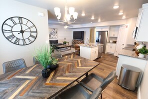 A modern kitchen with a central island, a dining area with a large wooden table, and a living area with a sofa and a large wall clock.