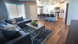 A modern living room with a sofa, coffee table, and patterned rug. A kitchen with white cabinets and stainless steel appliances is visible in the background.