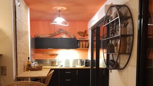 A narrow kitchen with a wooden table and two wicker chairs, a hanging light fixture, and a black-framed window with a decorative screen. - A charming setting with the sea in full view! (Pietrosella)