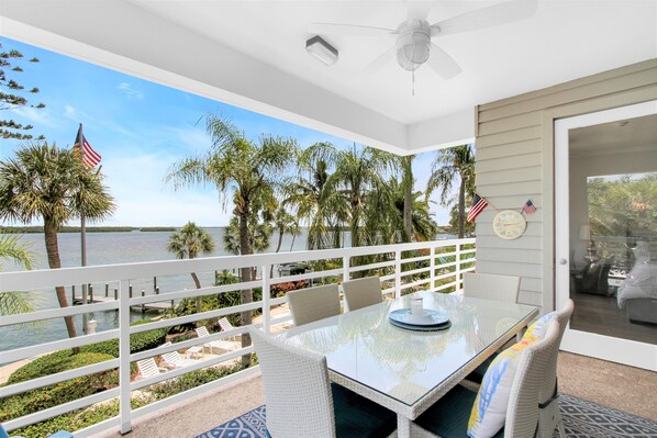 A balcony with a glass table, white wicker chairs, and a view of the water and palm trees.