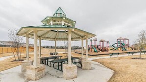 Um coreto com telhado verde, mesas de piquenique e equipamentos de playground ao fundo.