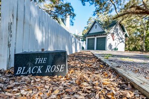 A sign reading "THE BLACK ROSE" on a stone slab, surrounded by fallen leaves, with a white fence and a building in the background.