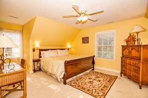 Ein Schlafzimmer mit einem Bett, einer Holzkommode, einem Schreibtisch mit Lampe, einem Deckenventilator und einem Fenster mit Jalousien.