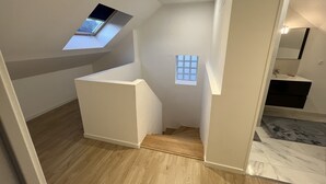 Ein modernes Interieur mit schrägverdeckter Decke, einem Oberlicht, einem kleinen Fenster und einer Treppe, die in ein unteres Geschoss führt.