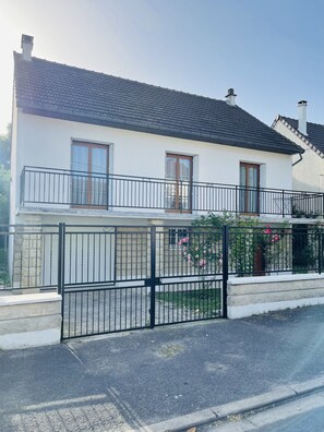 A two-story house with a balcony, a gated entrance, and a garden with pink flowers.
