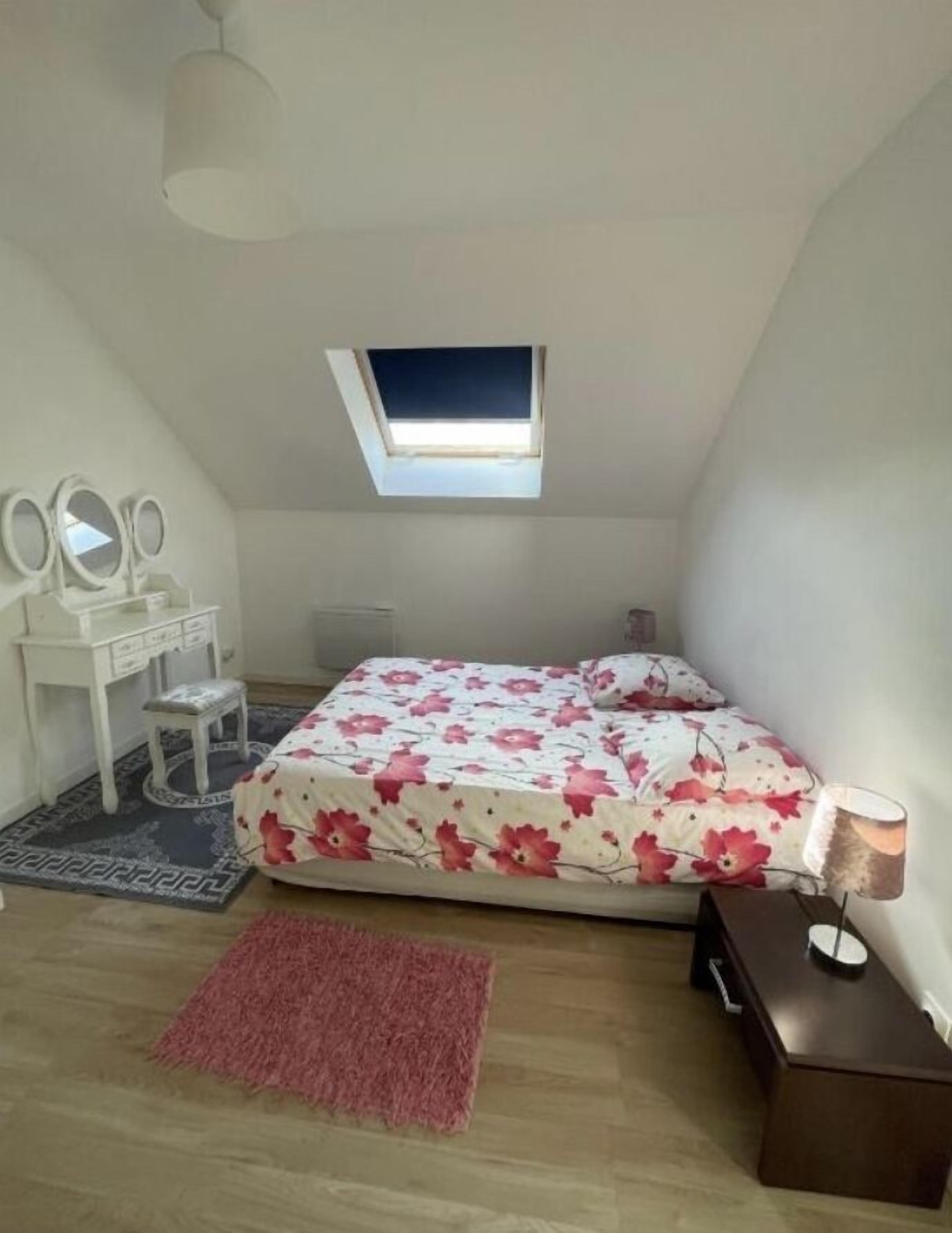 Ein Schlafzimmer mit Dachfenster, einem Bett mit floralem Überwurf, einem Schminktisch mit Spiegeln, einem kleinen Hocker, einem Teppich und einem Nachttisch mit Lampe.