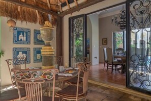 A dining area with a rustic thatched ceiling, a large decorative urn on the table, and a view through a glass door to an outdoor patio. - Casa La Perlita - Large Luxury Colonial in Centro - Garden, Gazebo, Big Pool ❤️ (Mérida)
