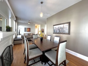 A dining area with a long wooden table, six chairs, a vase with flowers, and a cityscape painting on the wall.