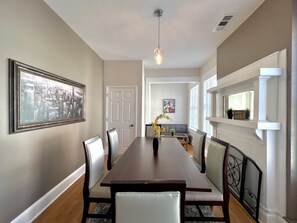 A dining room with a long wooden table, cushioned chairs, a hanging light fixture, and a cityscape painting on the wall. - ⌂ Elevated 2bed 1.5 Bath in a Historic 1913 Building ⌂ (Atlanta)