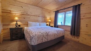 Un dormitorio de cabaña de madera con una cama, dos mesitas de noche y un ventanal con cortinas.