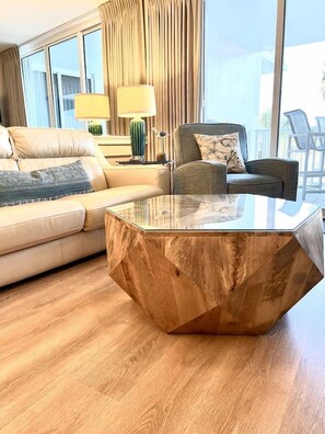A modern living room with a glass-top coffee table, a beige sofa, and a view of the outdoors through large windows. - Beach time! Destin West Condo Pelican 303 (Fort Walton Beach)