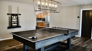 A ping pong table in a room with wooden flooring, a kitchen in the background, and a ceiling light fixture.