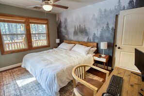 A bedroom with a bed, wooden headboard, nightstands, a desk with a computer, and a large window with a view of trees. - The Monarch (Pinetop-Lakeside)