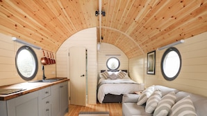 Un interior compacto de madera con cocina, cama y zona de estar, con ventanas redondas y techo curvo.