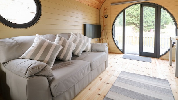 Una habitación acogedora con techo de madera, un sofá gris con cojines a rayas y un ventanal redondo con vistas al exterior.