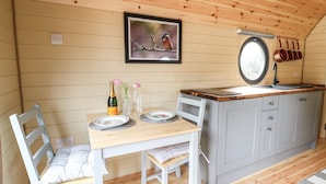 Un acogedor interior de cabaña con una mesa de comedor de madera para dos comensales, una pequeña cocina con un fregadero y un ventanal que deja entrar luz natural.