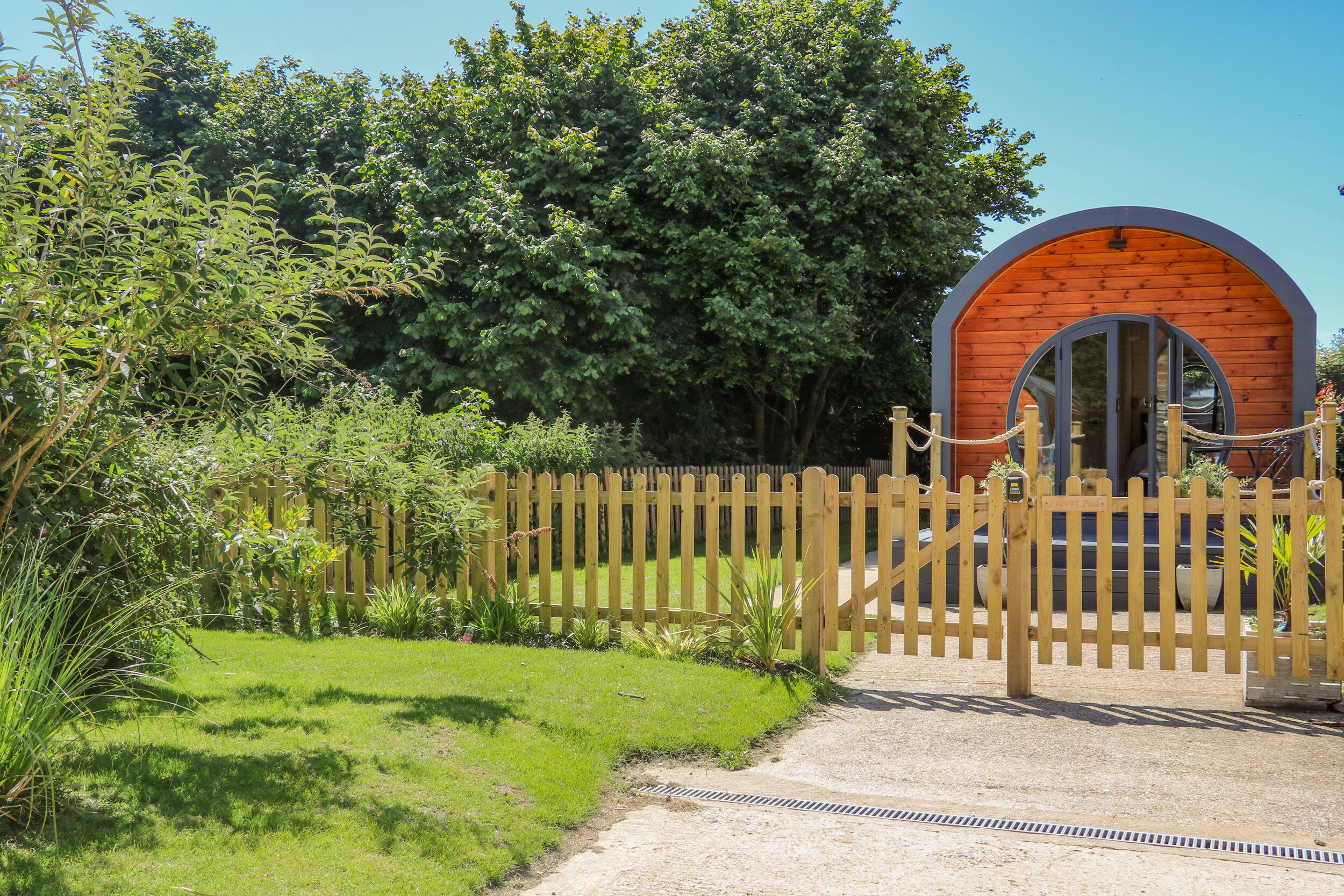 A wooden cabin with a circular arch entrance, surrounded by a wooden fence and lush greenery.