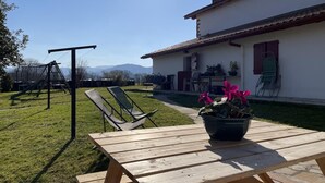 Une table de pique-nique en bois, avec une plante en pot au premier plan, une balançoire et une maison avec un toit rouge en arrière-plan.