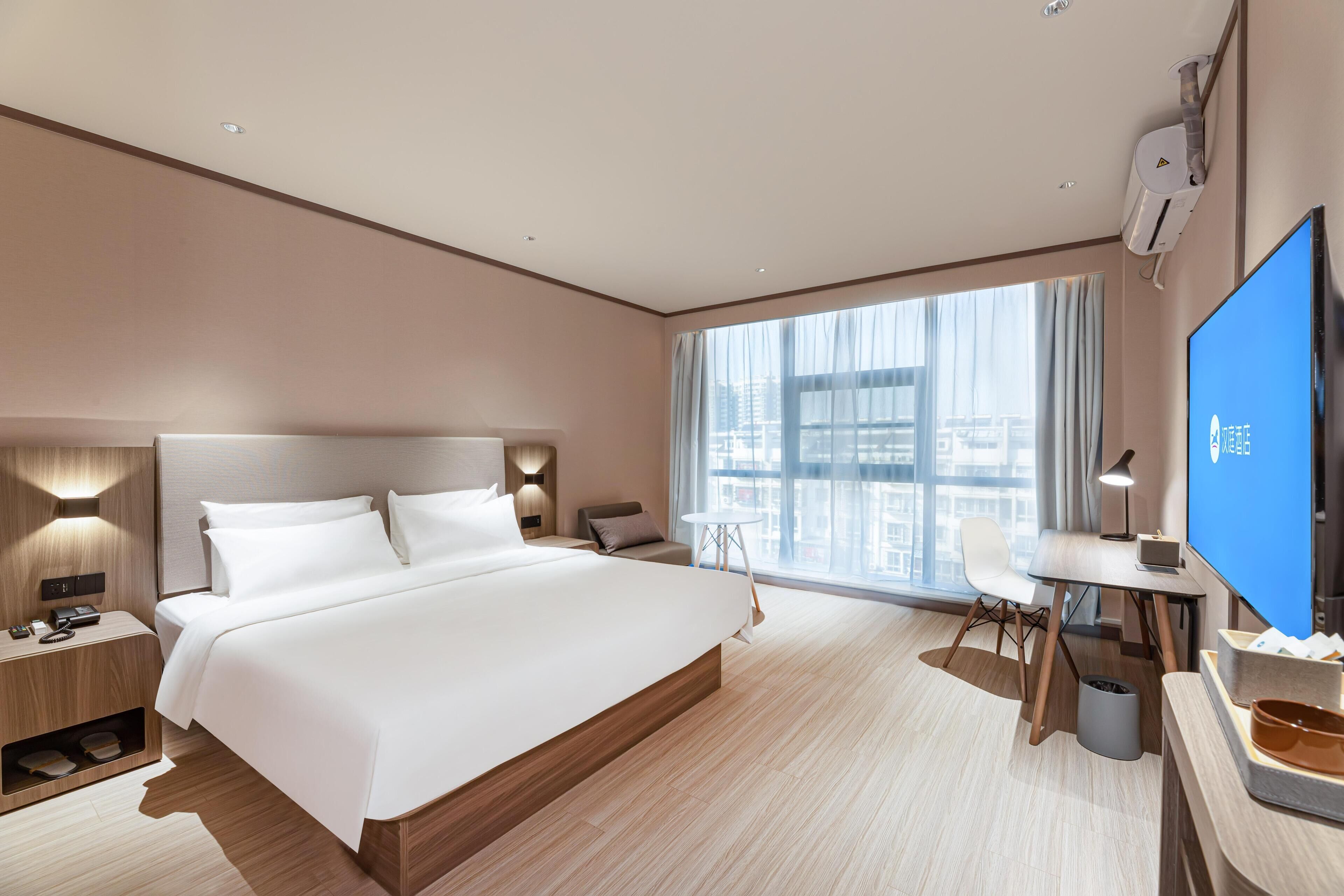 A modern hotel room with a large bed, a desk, a chair, a TV, and a window with a city view.