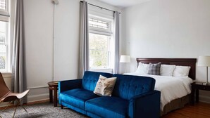 Camera d'albergo con divano blu, letto con biancheria bianca, testiera in legno, comodino con lampada, una poltrona e una finestra con tende.