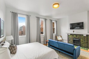 A hotel room with a bed, a blue sofa, a television, a fireplace, and large windows. - Chic Pet-Friendly Studio with Hotel-Like Amenities (Toledo)